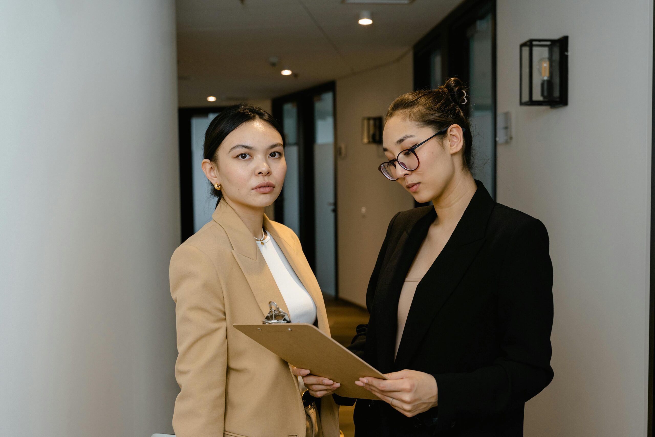 Two businesswomen in formal attire discuss work in a modern office corridor.